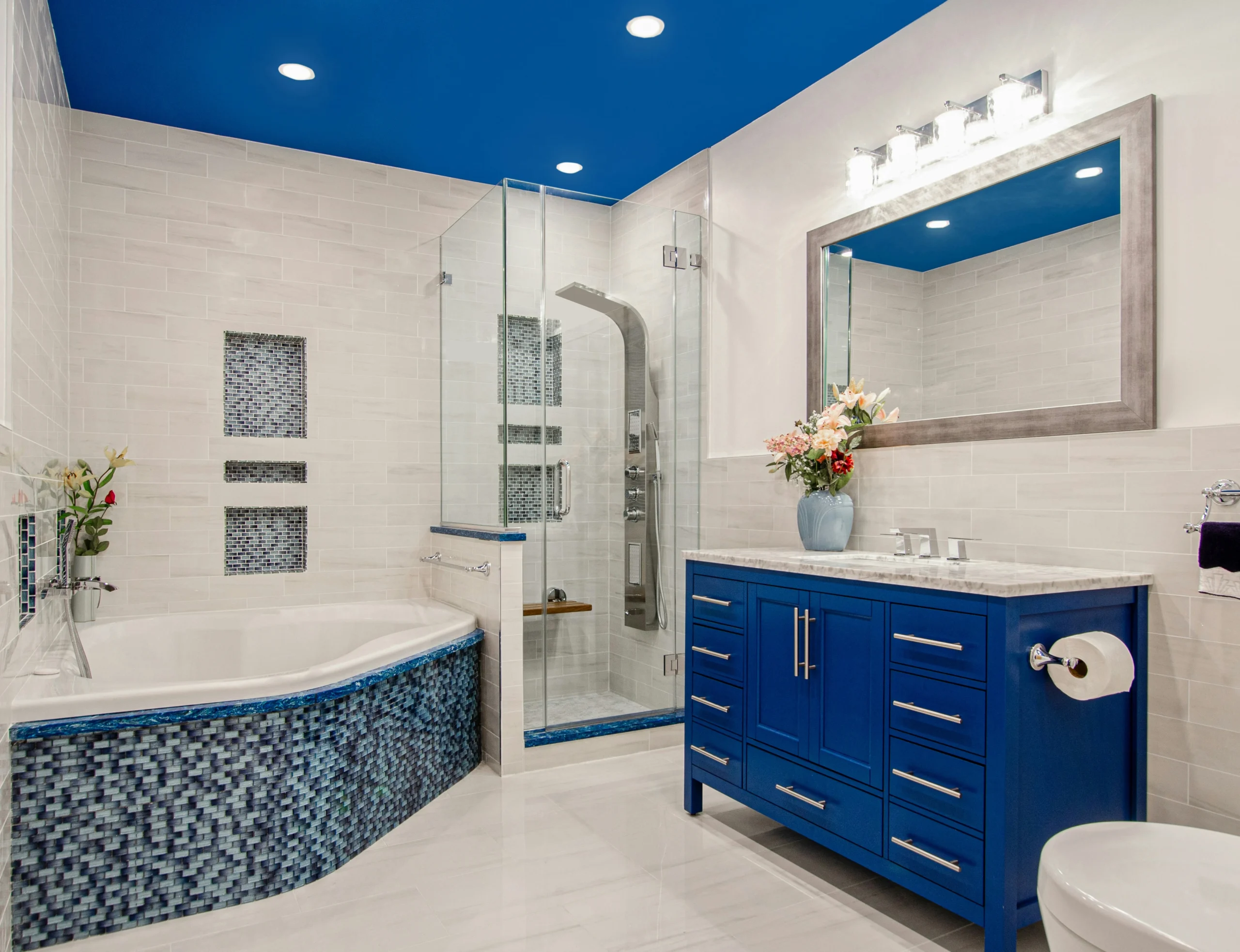 Modern bathroom renovation featuring a blue vanity, glass-enclosed shower, mosaic tile accents, corner bathtub, and bold blue ceiling.
