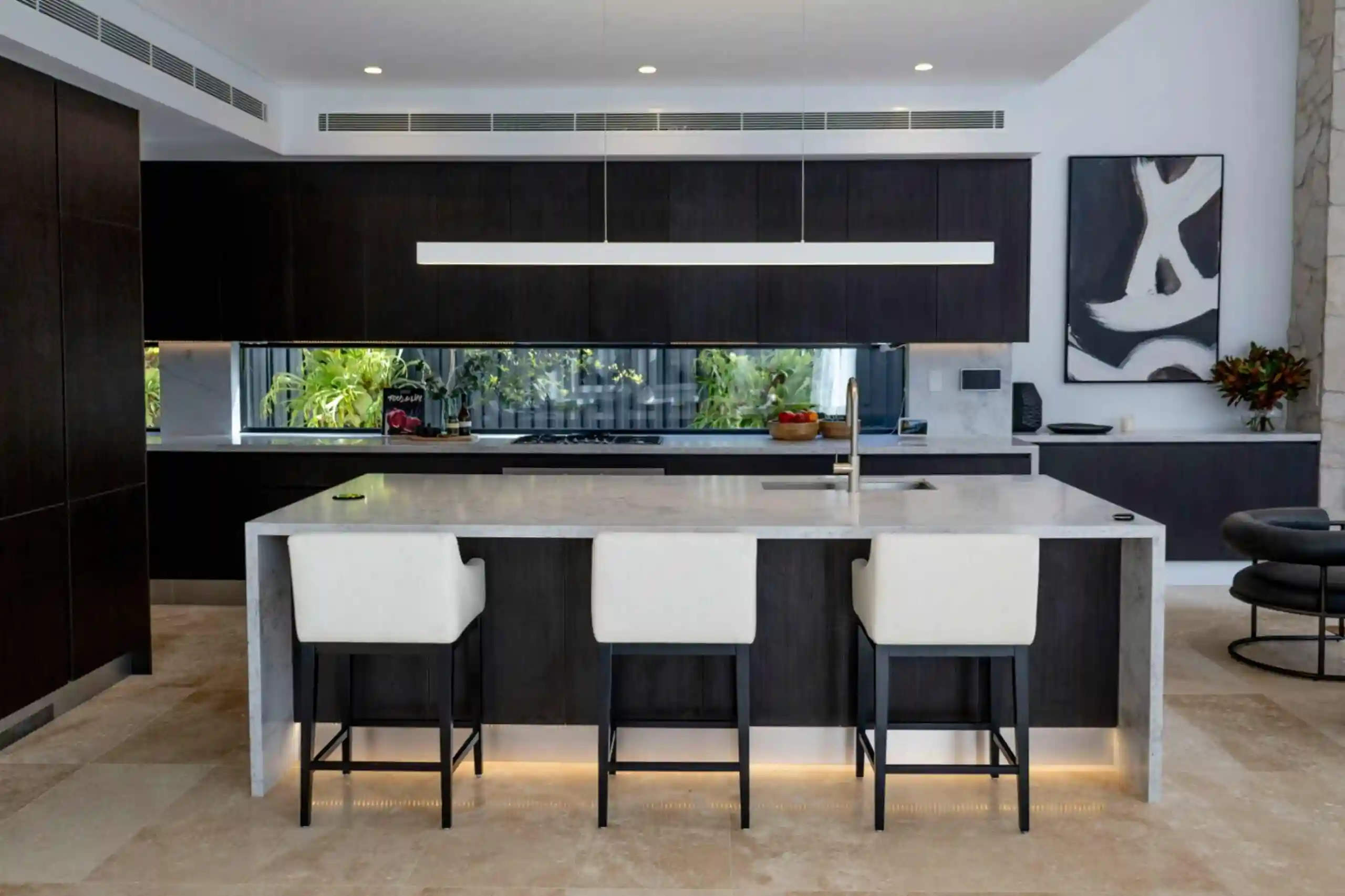 Contemporary kitchen with dark cabinetry, marble island countertop, white bar stools, under-cabinet lighting, and a window backsplash showcasing greenery.
